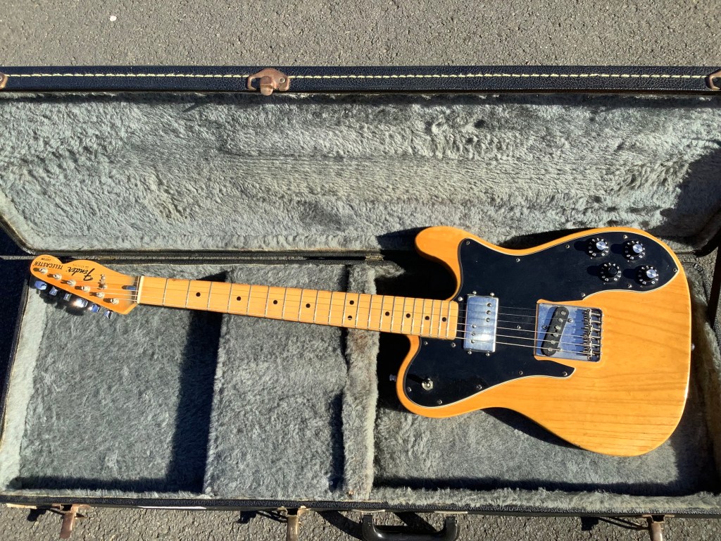A natural wood Fender Telecaster Custom Deluxe guitar resting in a hard case, featuring a black pickguard, two humbuckers, and control knobs.